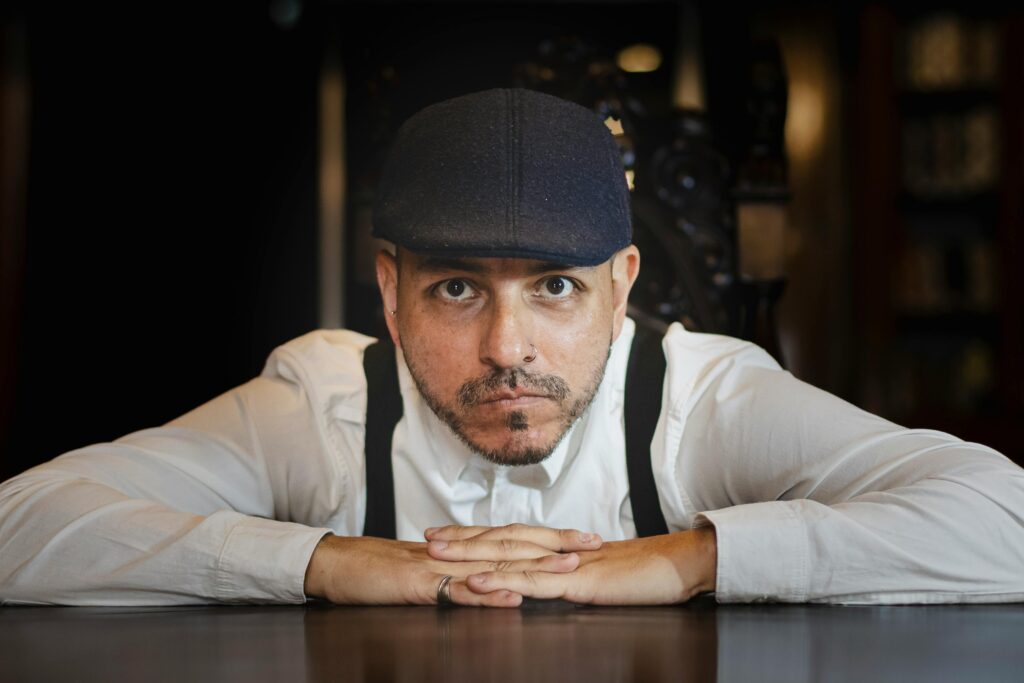 Close-up portrait of a man wearing a cap and white shirt, leaning on a table in a dimly lit room.